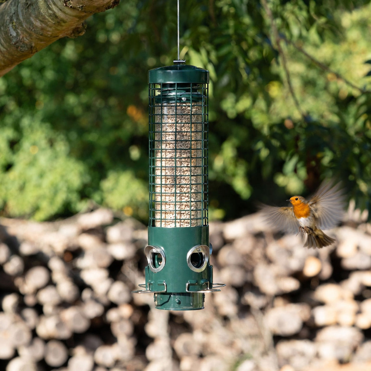 A green cylindrical bird feeder hanging from a tree branch, with a small orange-breasted bird flying nearby against a blurred outdoor background of green foliage and stacked logs.